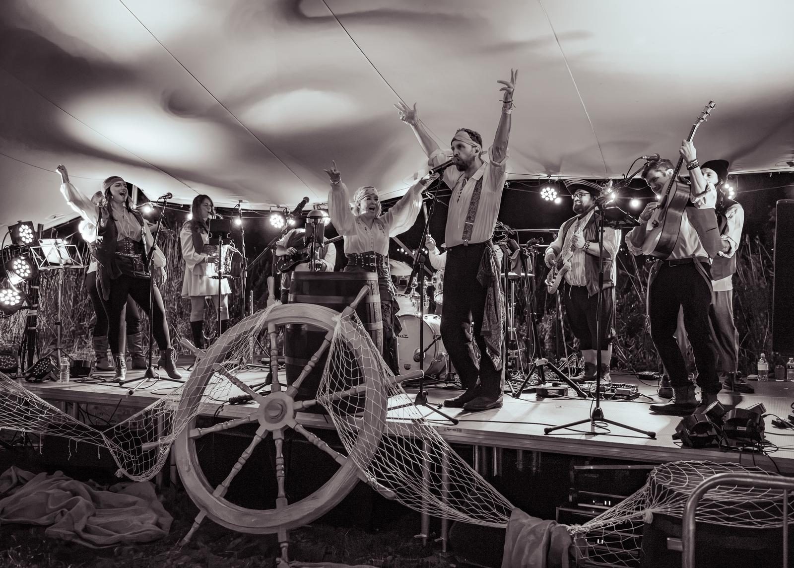 The full band on stage under the big top with arms raised, a ship’s wheel and fishing nets at the front of the stage