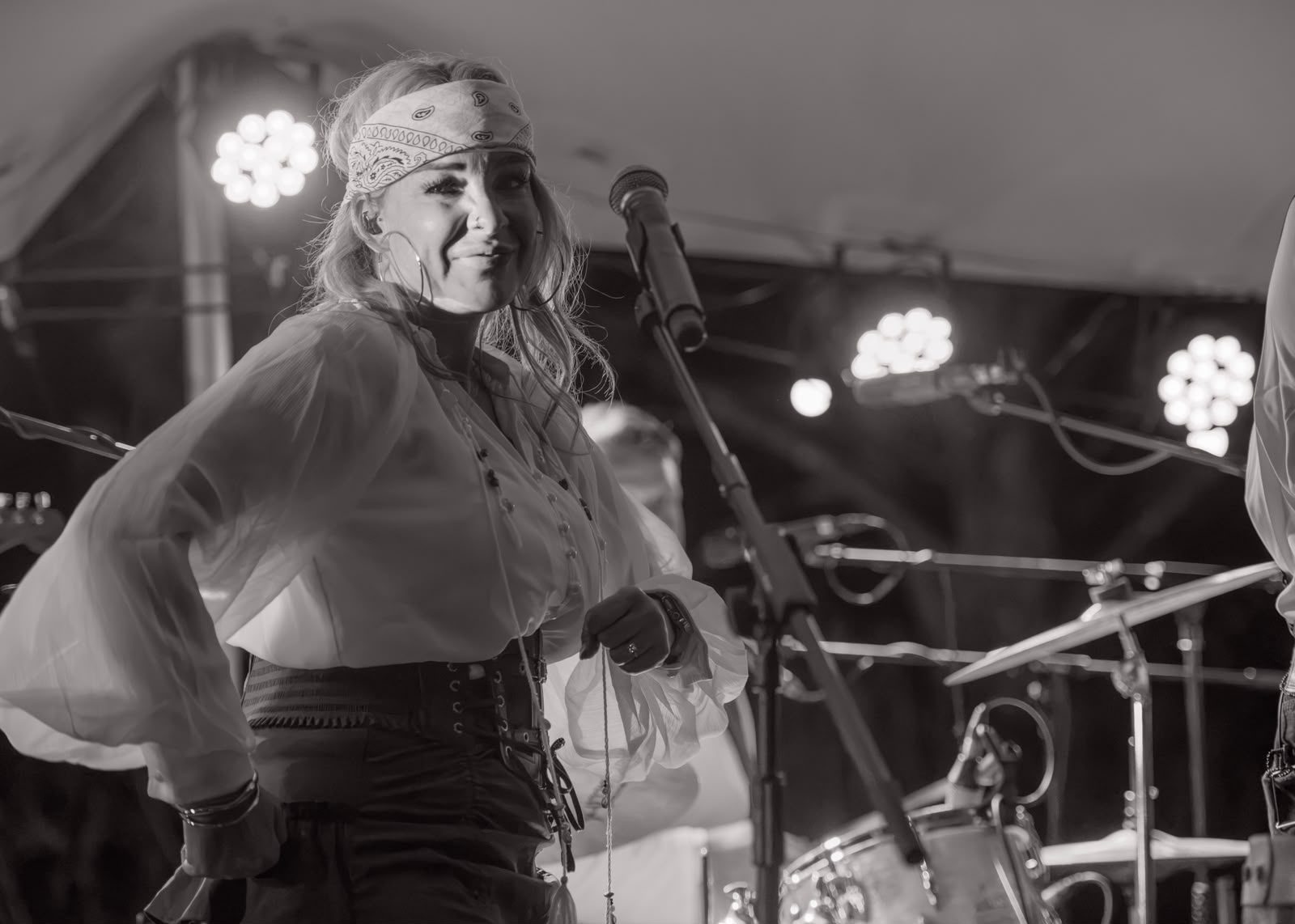 A singer in a white blouse and paisley bandana grinning beside the microphone, drums behind her